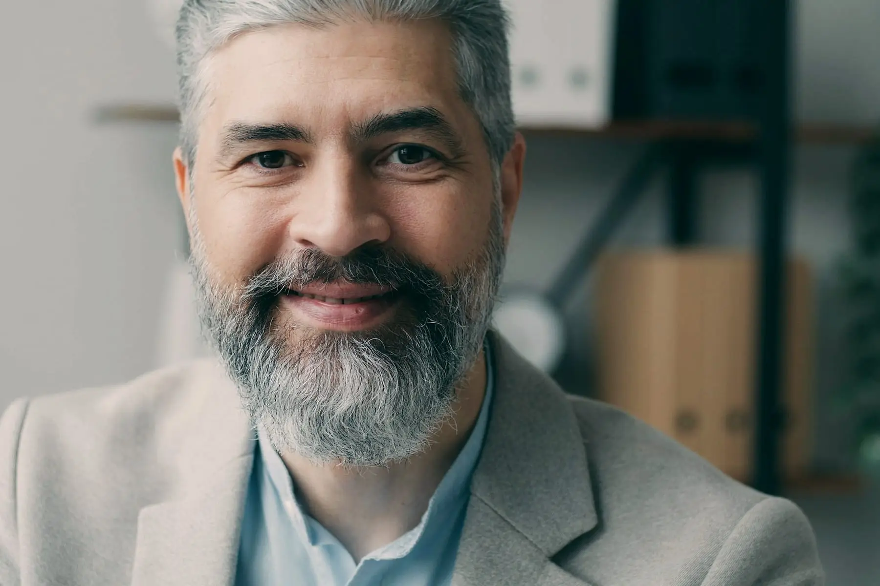 Homme mûr à la barbe grise, en veste beige, souriant dans son bureau