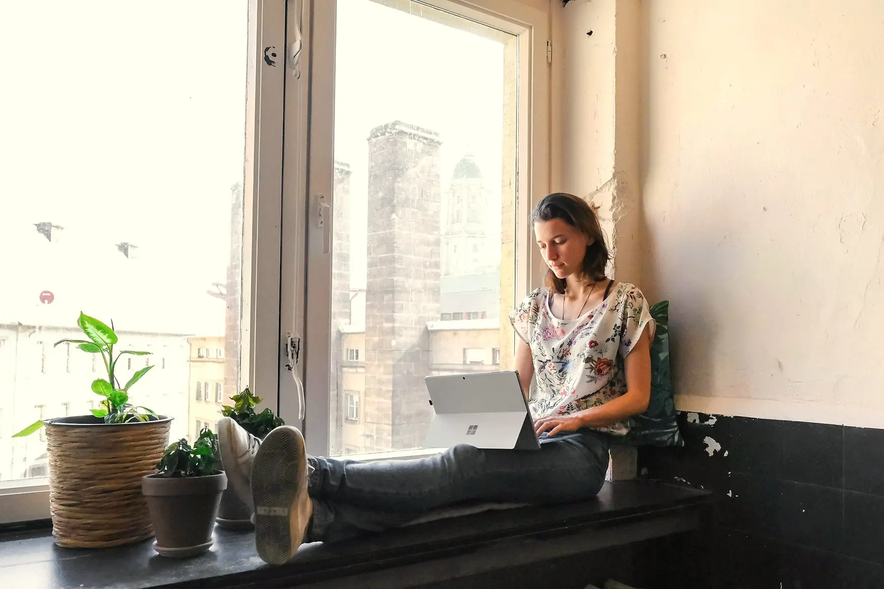Femme assise sur un rebord de fenêtre avec son ordinateur, entourée de plantes
